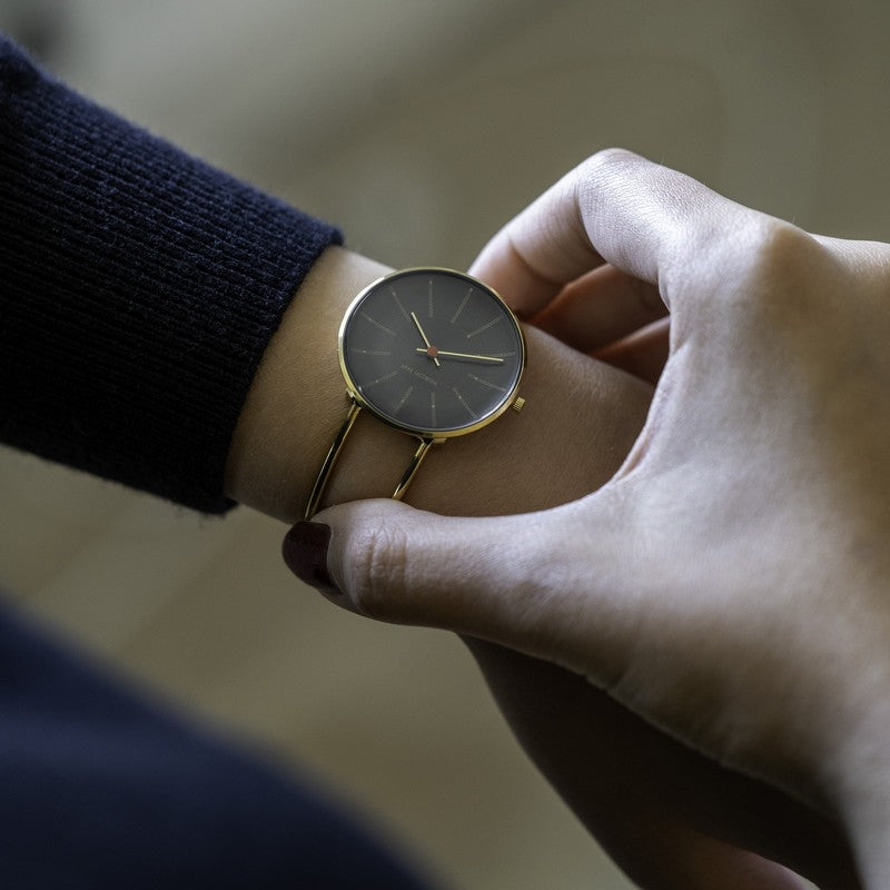 Arne Jacobsen Bankers jubilæumsur i rustfrit stål med grå skive og guld bangle, set fra siden